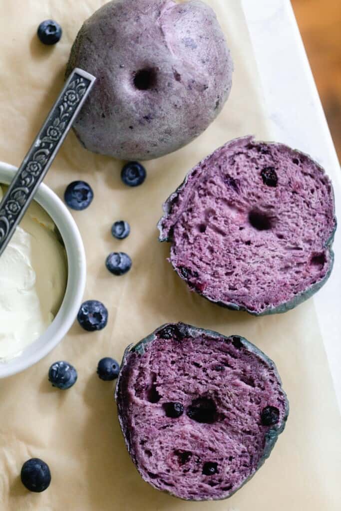 blueberry sourdough bagel sliced in half  and another whole bagel on parchment paper with blueberries sprinkled around. A container of cream cheese with a knife is to the left