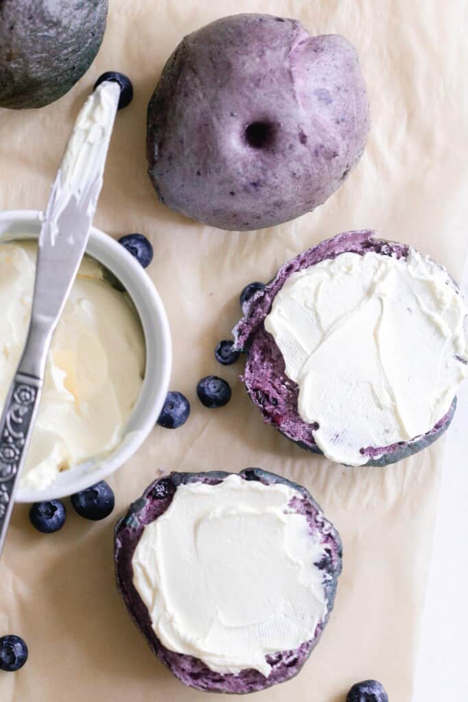 overhead photo of a sourdough blueberry bagel sliced in half and topped with cream cheese on parchment paper. Another bagel and a container of cream cheese to the left of sliced bagel
