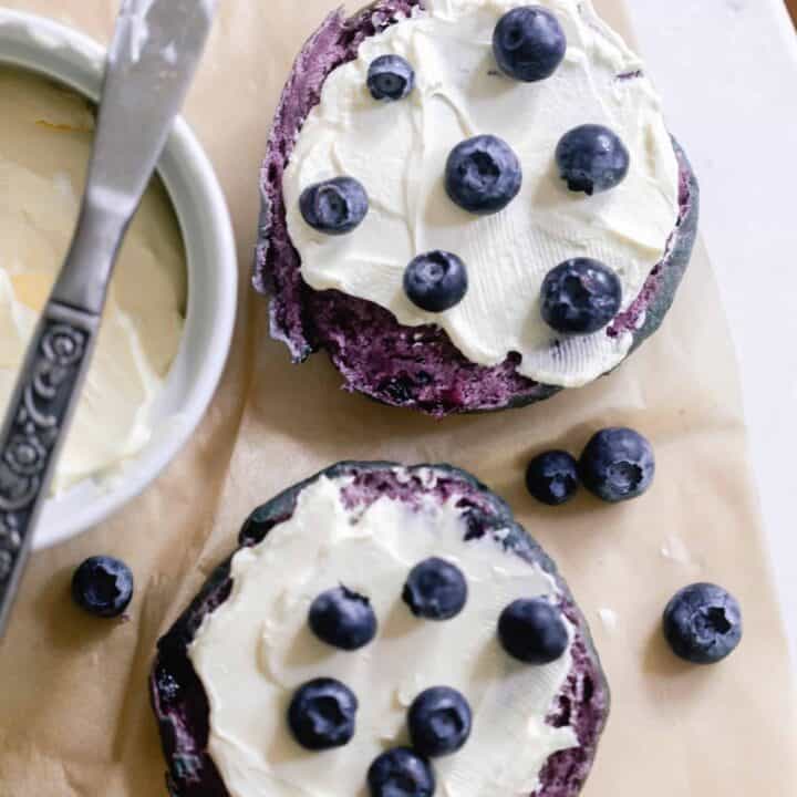overhead photo of a sourdough blueberry bagel sliced in half and topped with cream cheese topped with blueberries. The bagels are on parchment paper with a cream cheese in a white container and topped with a knife