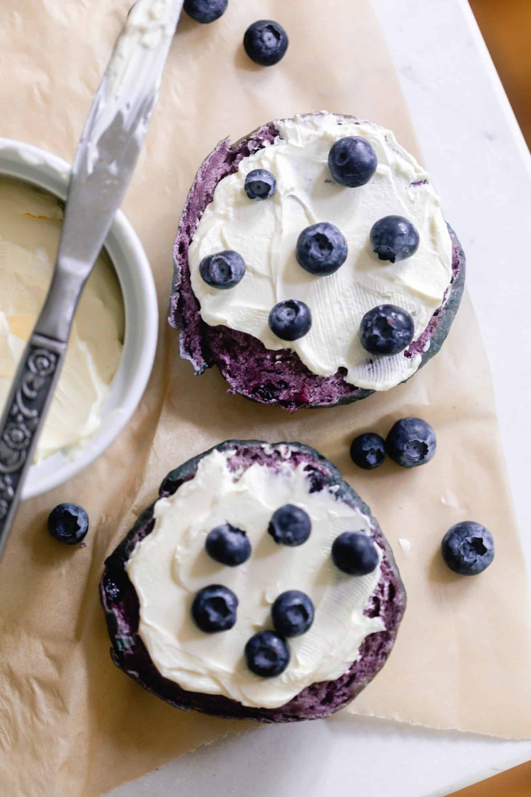 overhead photo of a sourdough blueberry bagel sliced in half and topped with cream cheese topped with blueberries. The bagels are on parchment paper with a cream cheese in a white container and topped with a knife