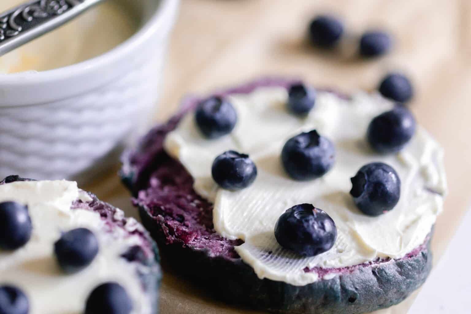 close up picture of a sliced blueberry sourdough bagel with cream cheese spread and topped with blueberries.