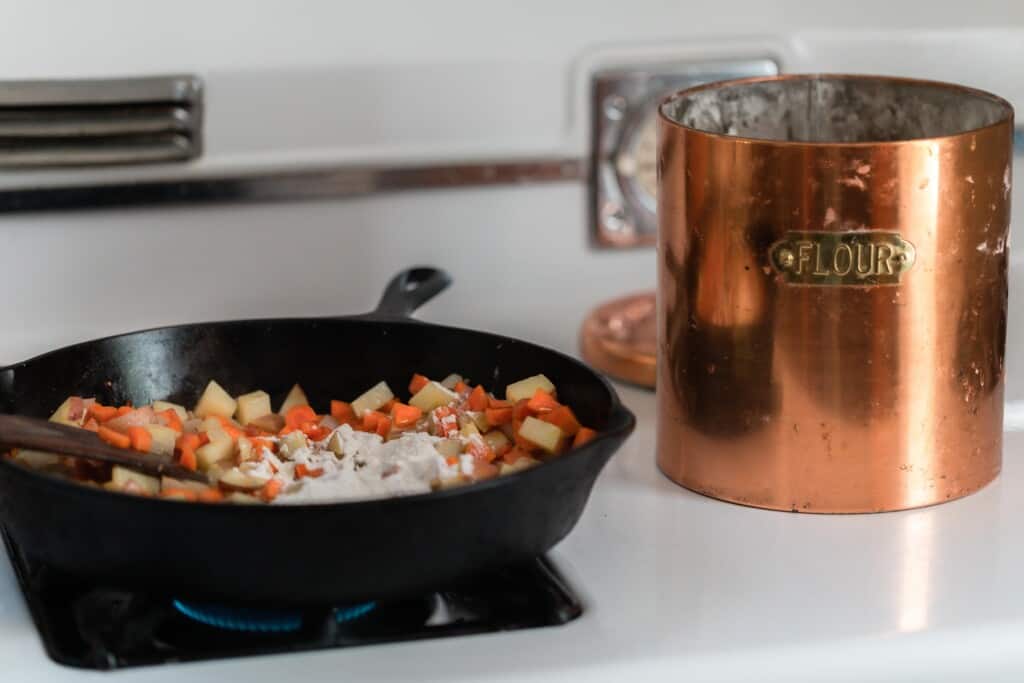 carrots, potatoes, onions sautéing in a cast iron skillet, with flour sprinkled over. A copper canister of flour to the right.