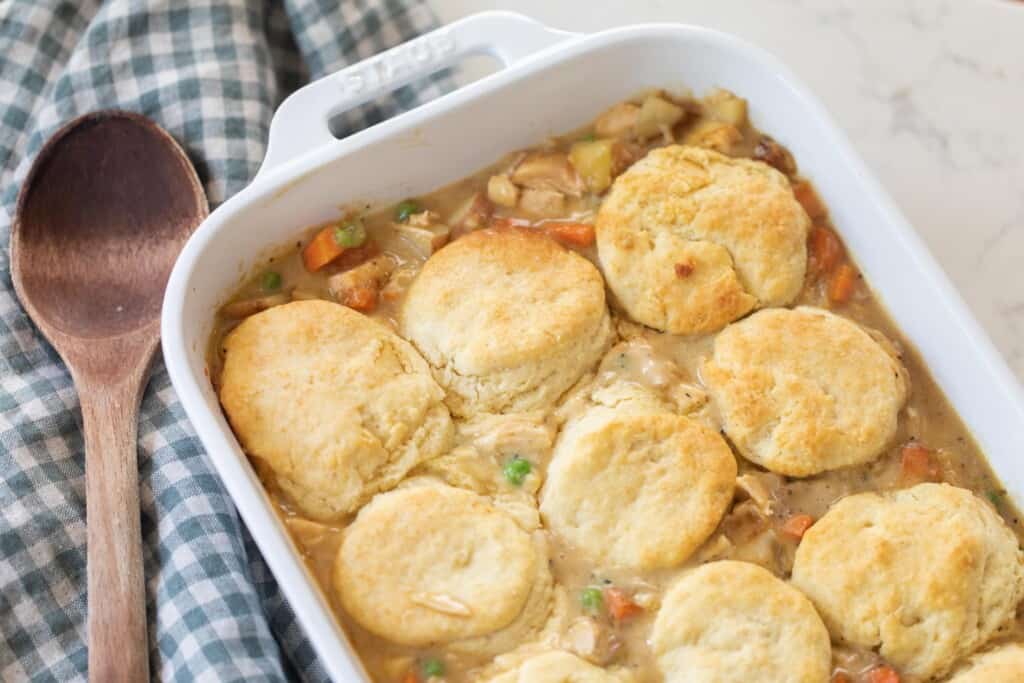 chicken pot pie topped with sourdough biscuits in a white baking dish on a white and blue checked towel with a wooden spoon to the left