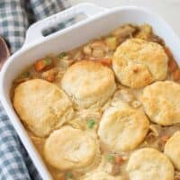 chicken pot pie topped with sourdough biscuits in a white baking dish on a white and blue checked towel with a wooden spoon to the left