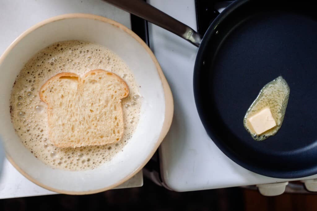 brioche in a custard mixture in a bowl next to a cast iron skillet with butter melting