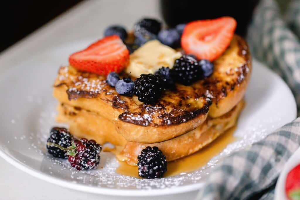 close up picture of two pieces of sourdough French toast topped with fruit, butter, powder sugar, and maple syrup on a white plate