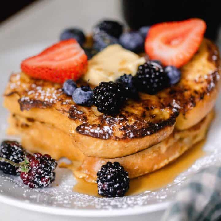 close up picture of two pieces of sourdough French toast topped with fruit, butter, powder sugar, and maple syrup on a white plate