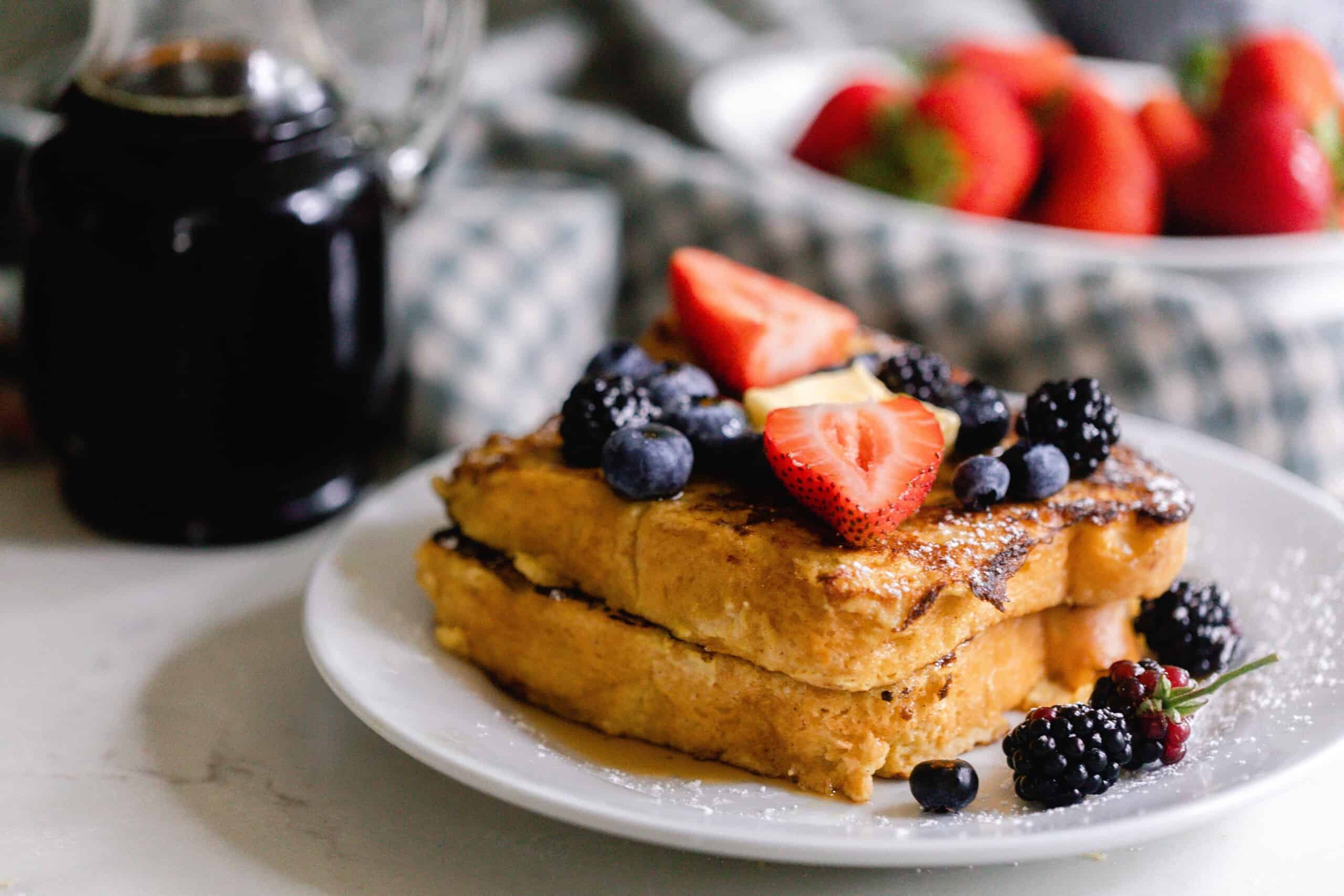 two pieces of sourdough French toast on a white plate and topped with fresh fruit. Maple syrup and strawberries in a bowl are in the background