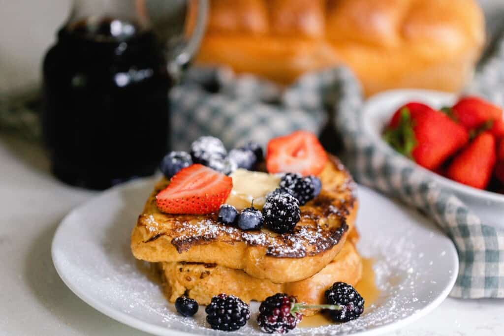 sourdough French toast topped with strawberries, blueberries, a pat of butter, and powdered sugar on a white plate with maple syrup and blue checked towel and a bowl of strawberries in the background