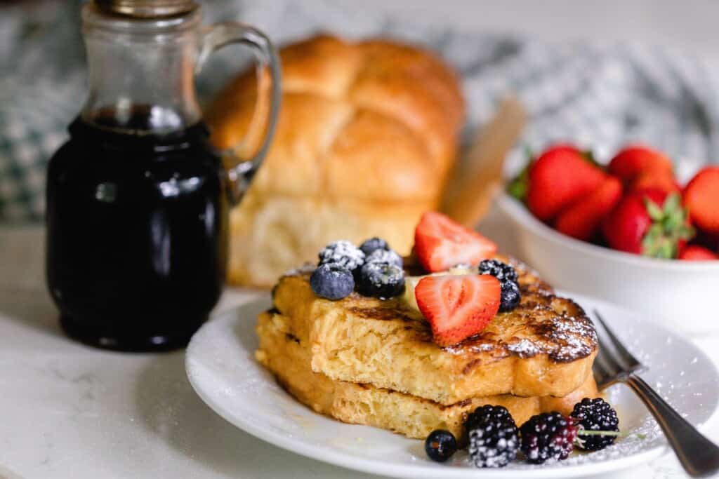 sourdough French toast topped with berries on a white plate with a bottle of maple syrup, a loaf of brioche, and a bowl of strawberries in the background