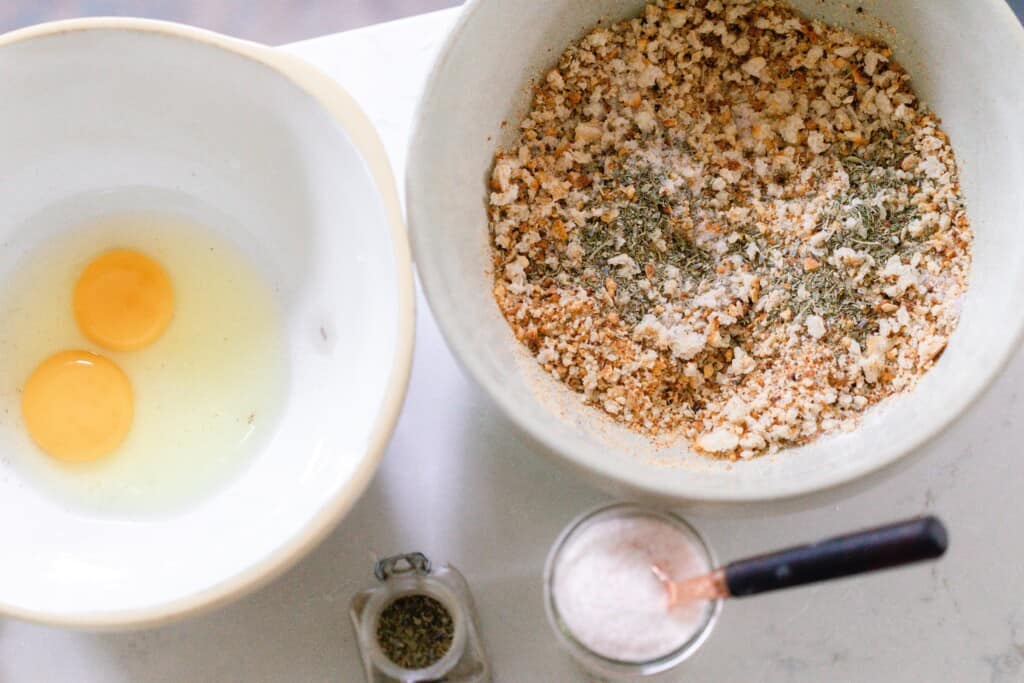 a bowl with eggs and another bowl with bread crumbs and herbs to the right