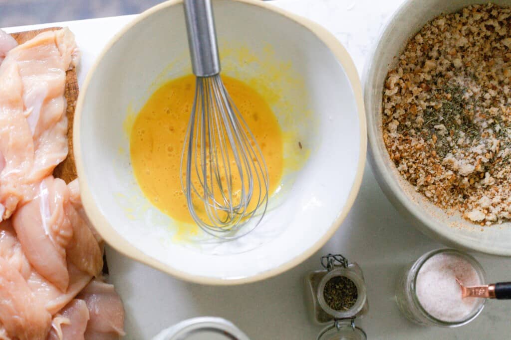 sliced chicken, a bowl of eggs with a whisk, and another bowl of bread crumbs on a white countertop