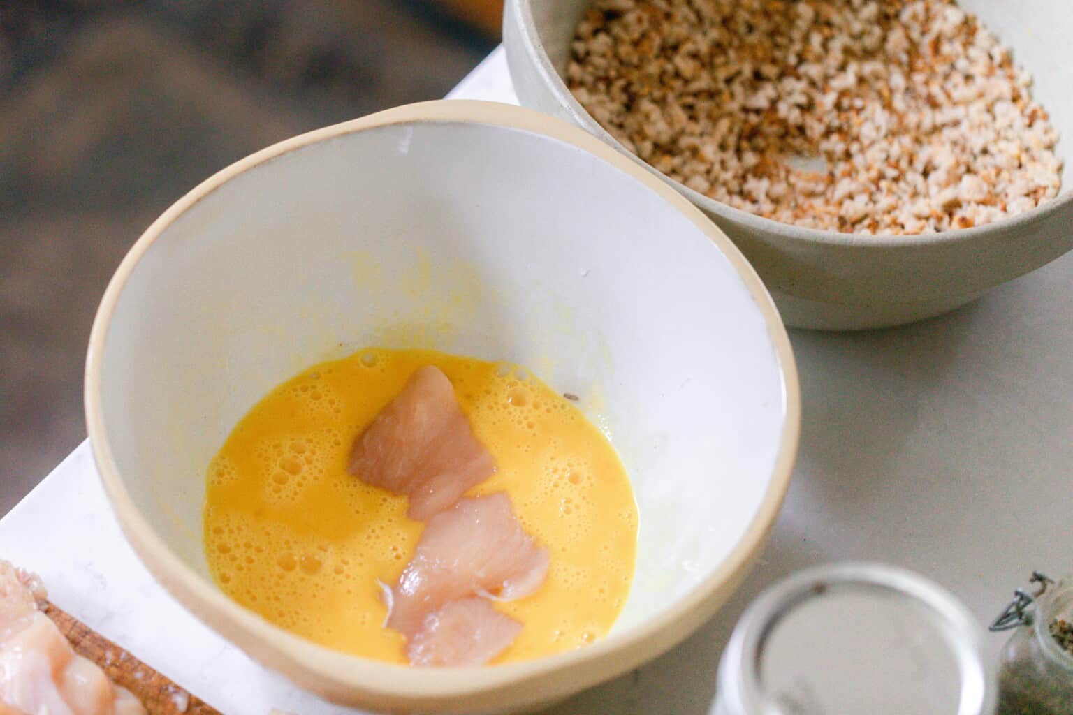 adding chicken to eggs in a bowl with another bowl of breadcrumbs in the background
