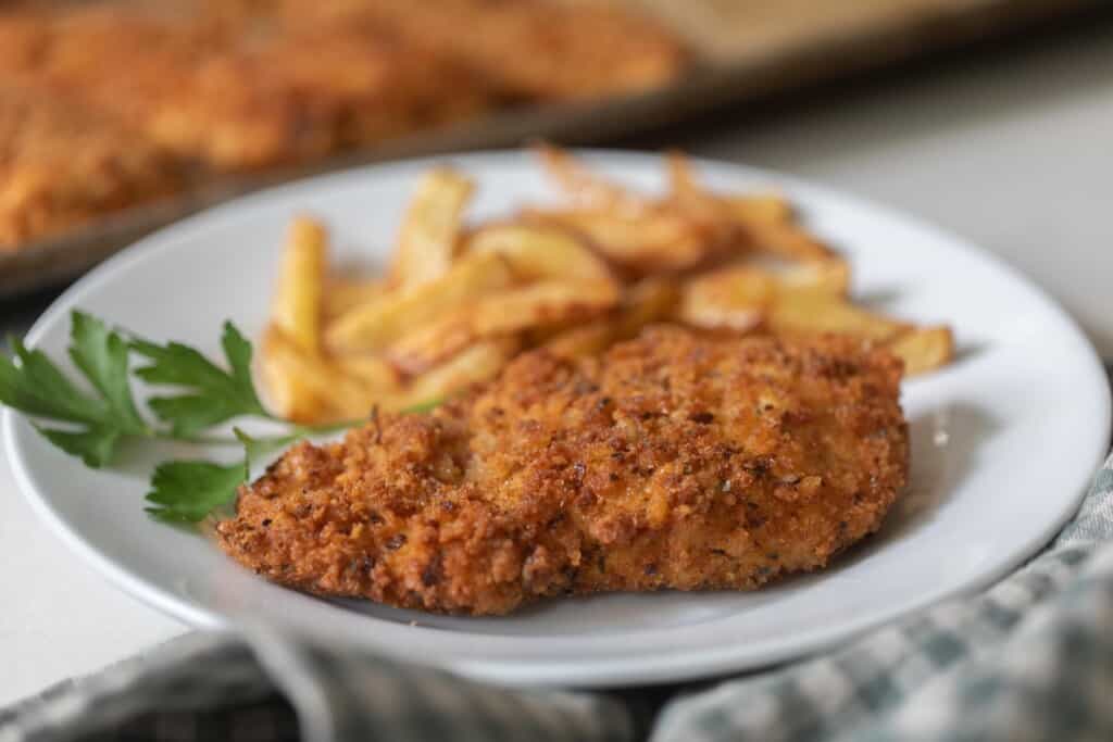 sourdough fried chicken on a white plate with French fries behind the chicken with a sprig of parsley to the left