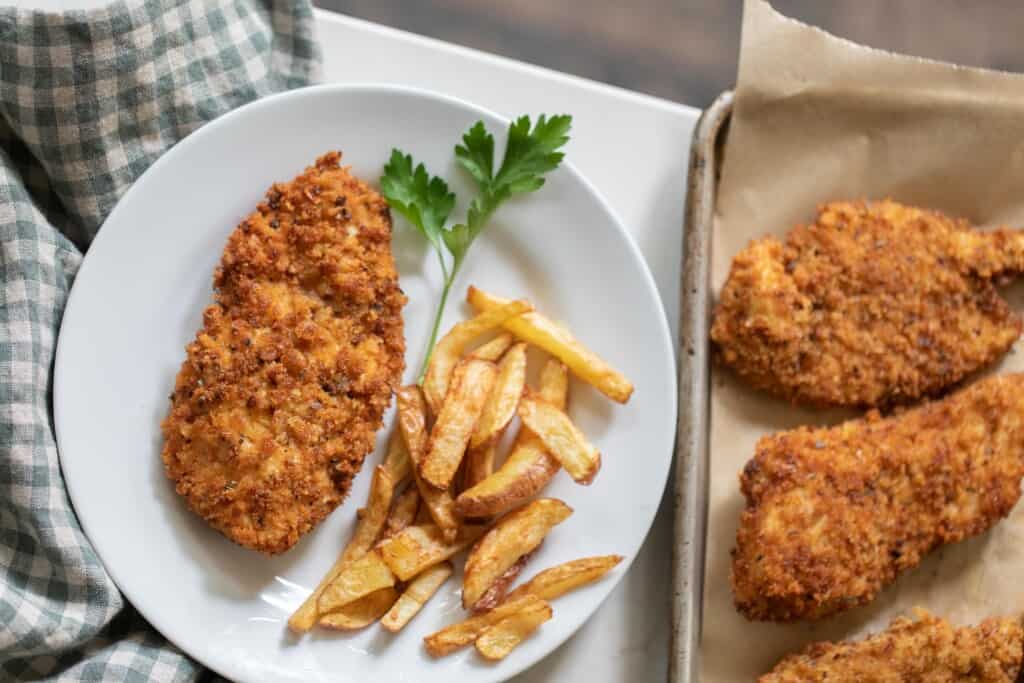 a plate of sourdough fried chicken and homemade fries with a sprig of parsley on a white plate with a baking sheet of more fried chicken to the right