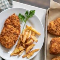 a plate of sourdough fried chicken and homemade fries with a sprig of parsley on a white plate with a baking sheet of more fried chicken to the right