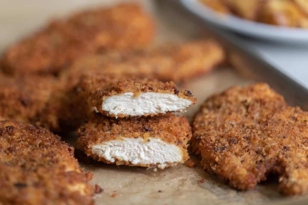 a parchment lined baking sheet full of sourdough fried chicken. One chicken breast is cutting half and stacked on top of each other