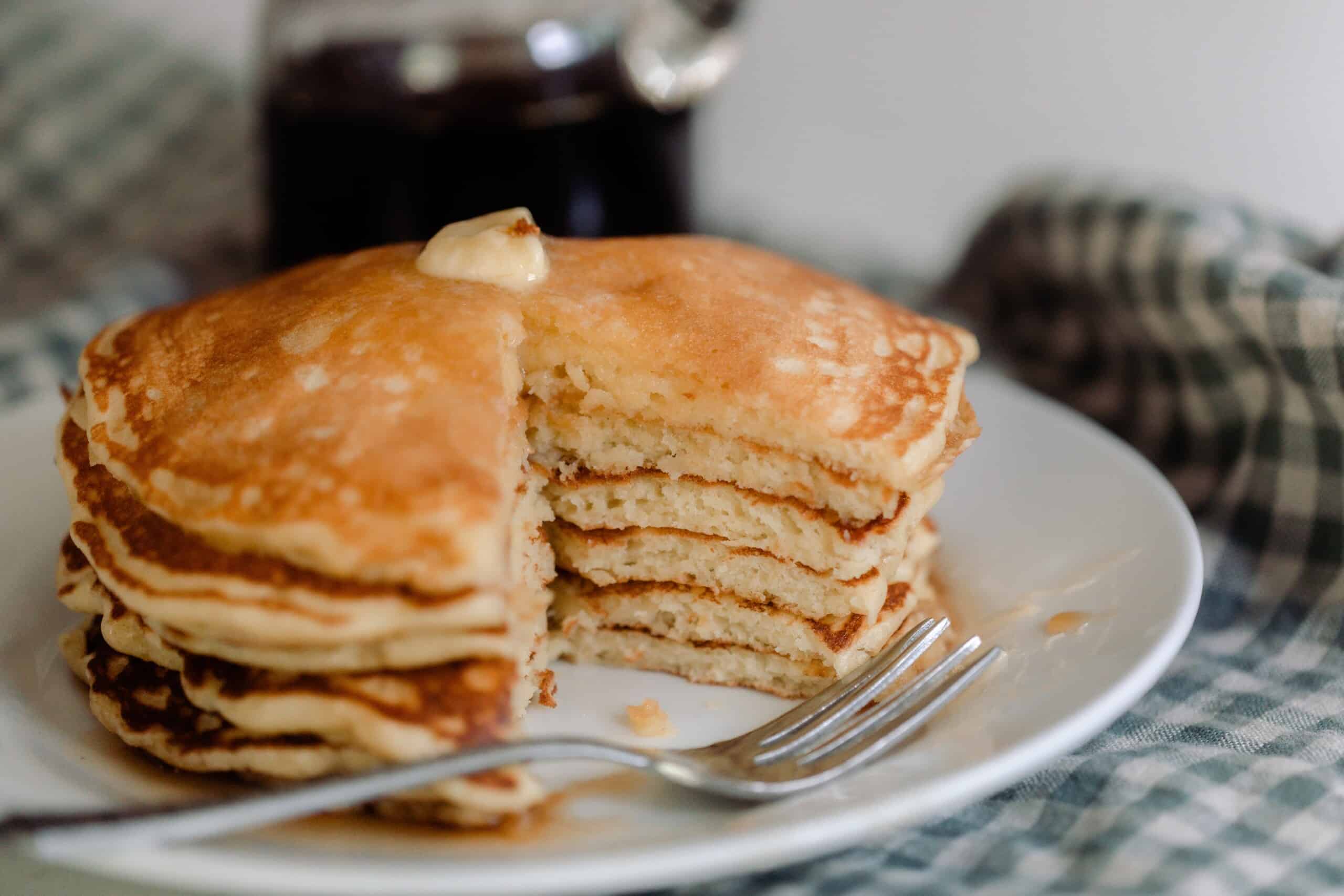 A stack of sourdough pancakes with a bite through all of the pancakes on a white plate with a fork beside it.