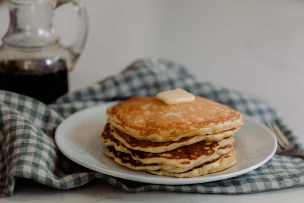 a white plate full of sourdough pancakes topped with butter on a white and blue checked towel. A bottle of maple syrup is in the background