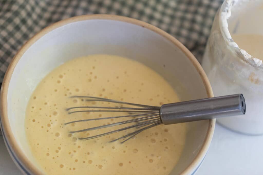 sourdough pancake batter in a stoneware bowl with a whisk in the batter. A glass jar of sourdough starter is to the right