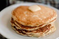 close up picture of sourdough pancakes stacked up 6 pancakes high with a pat of butter and maple syrup. The pancakes are on a white plate