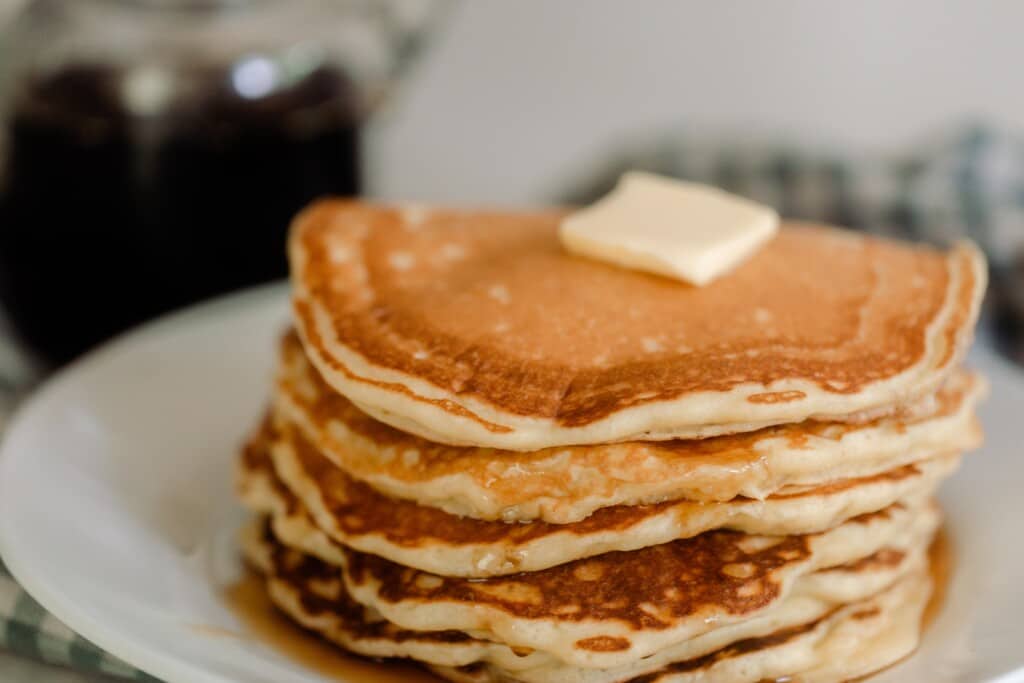 side view of a stack of sourdough pancakes with butter on a white plate with maple syrup in the background