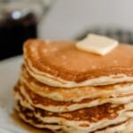 side view of a stack of sourdough pancakes with butter on a white plate with maple syrup in the background
