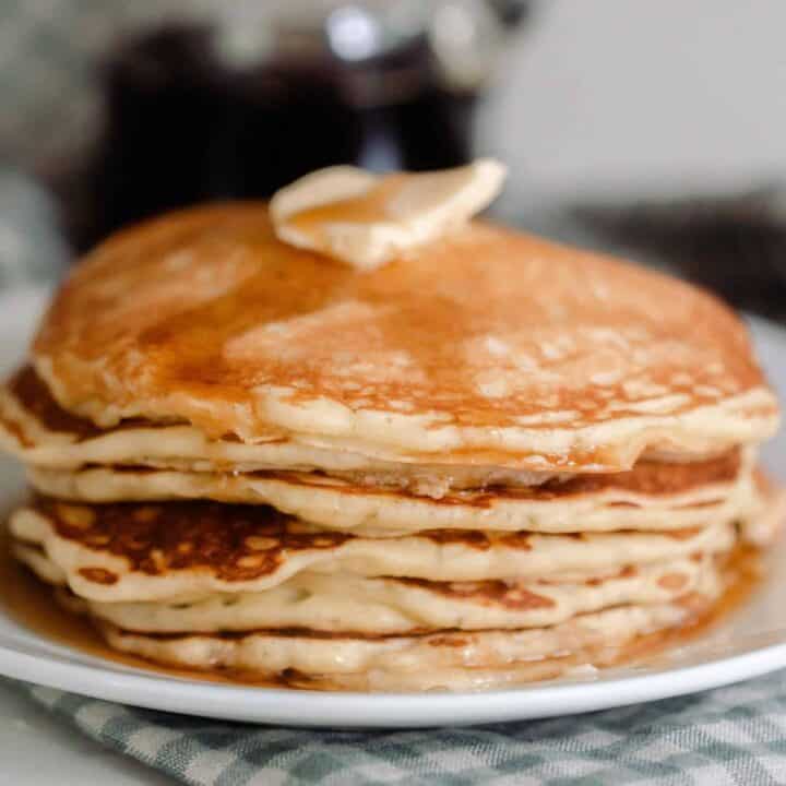 a stack of sourdough pancakes topped with a pat of butter and maple syrup on a white plate that rests on a blue and white checked towel