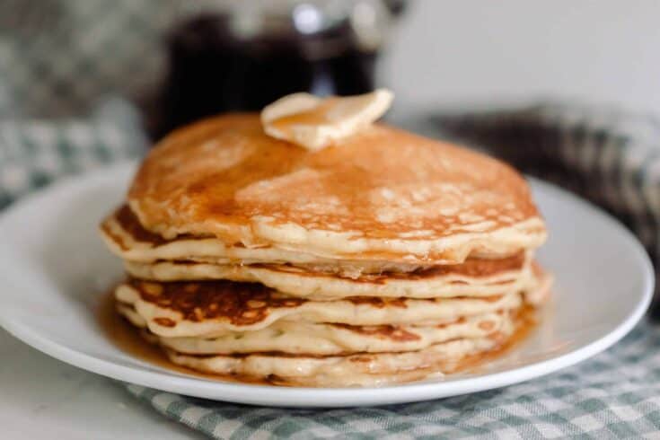 a stack of sourdough pancakes topped with a pat of butter and maple syrup on a white plate that rests on a blue and white checked towel