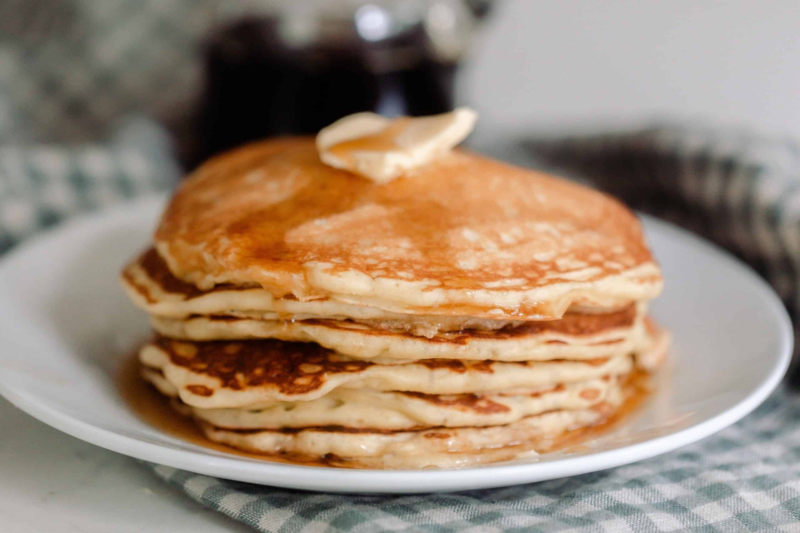 a stack of sourdough pancakes topped with a pat of butter and maple syrup on a white plate that rests on a blue and white checked towel