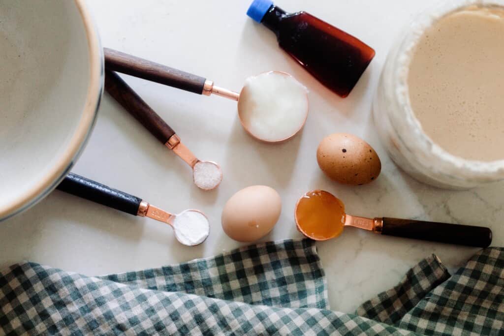 measuring cups and spoons of various ingredients, eggs, a jar of sourdough starter, and bowl on a white countertop with a blue and cream checked towel in the front