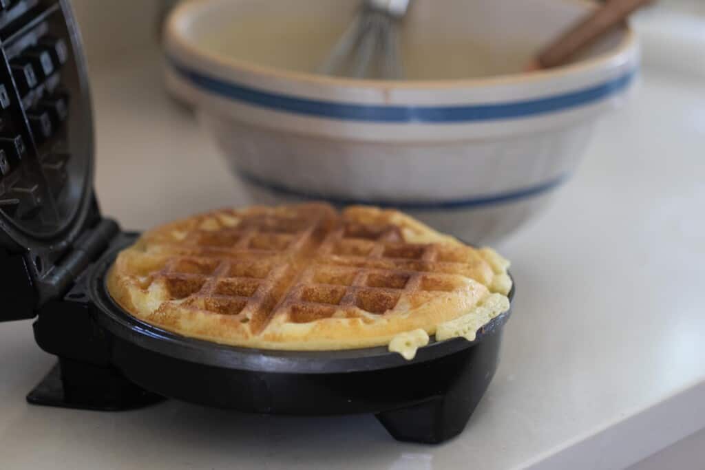 cooked waffle in a waffle iron with a bowl in the background