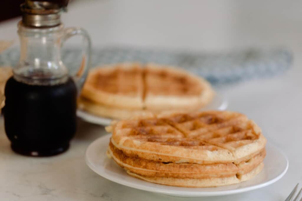 two stacks of sourdough waffles on white plates with a jar of maple syrup to the left