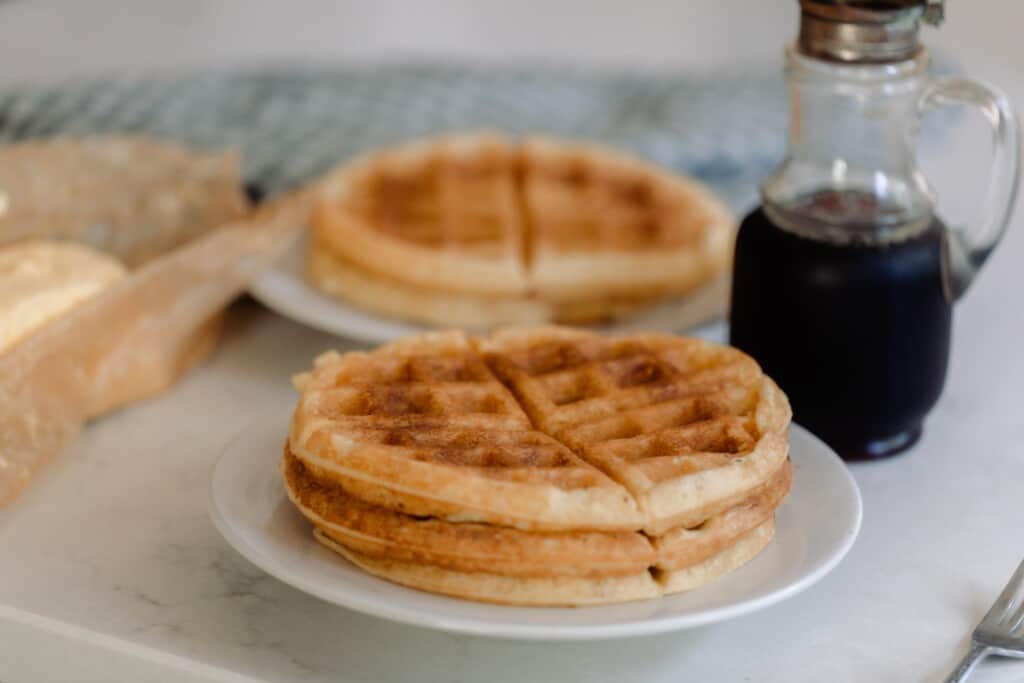 two stacks of waffles on plates on a white counter with a jar of maple syrup to the right