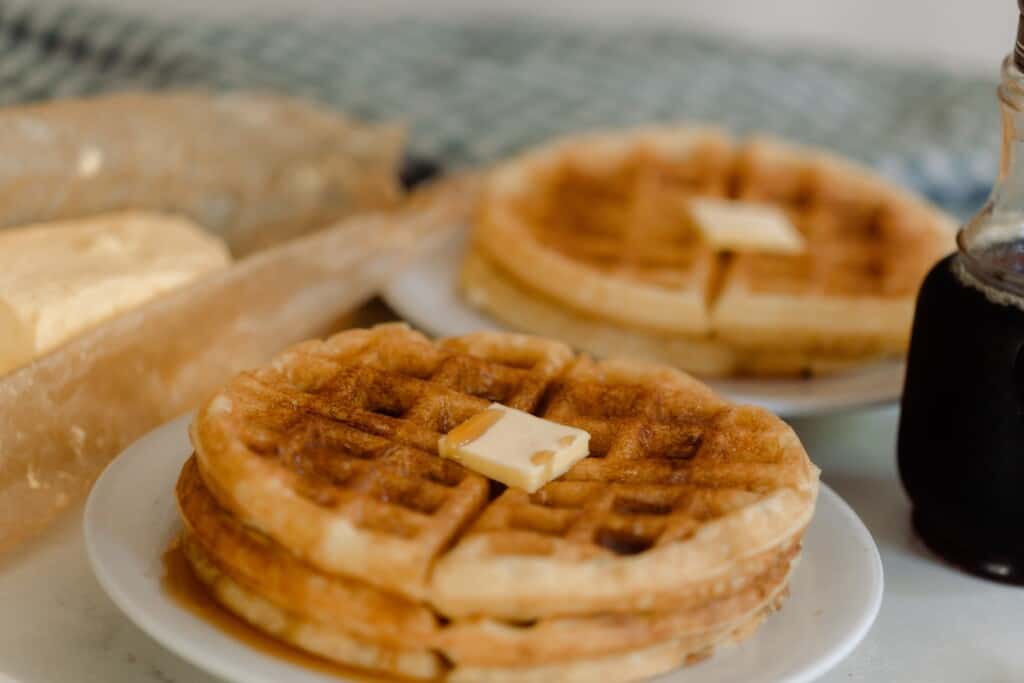 two plates stacked with sourdough waffles topped with butter and syrup. A bottle of syrup is to the left and butter on parchment paper is to the left