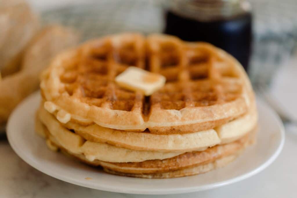 a stack of four sourdough waffles on a white plate. The waffles have a pat of butter and a jar of maple syrup in the background