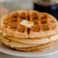 a stack of four sourdough waffles on a white plate. The waffles have a pat of butter and a jar of maple syrup in the background