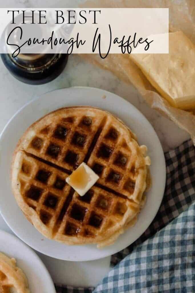 overhead photo of a crispy sourdough waffle topped with butter and maple syrup on a white plate on a white countertop. Butter on parchment paper and a jar of maple syrup is behind the waffles