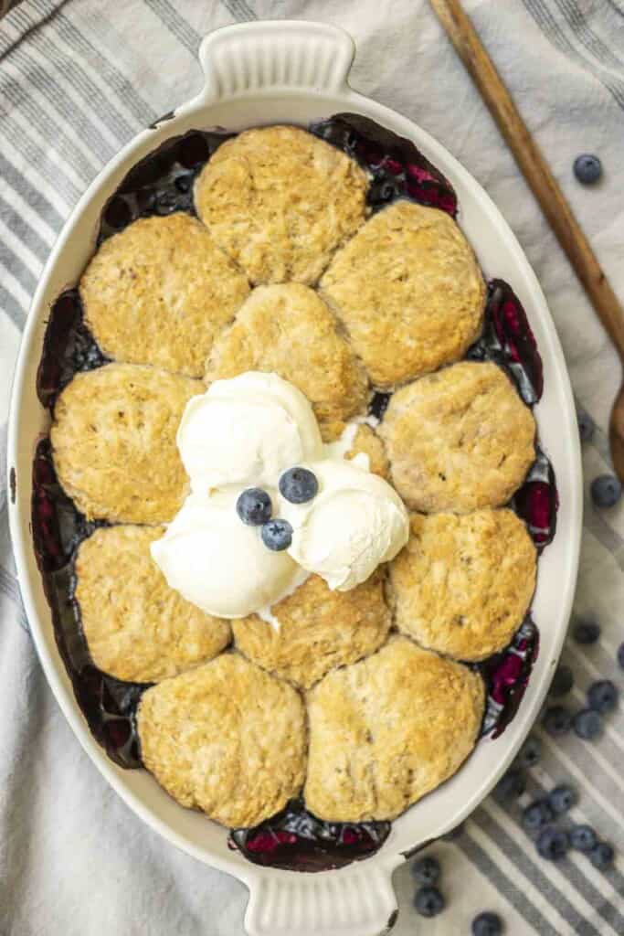 sourdough blueberry cobbler topped with ice cream and fresh blueberries in a white baking dish with fresh blueberries scattered around the dish