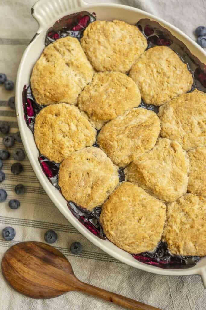 sourdough blueberry cobbler in a white baking dish on a white and blue towel with a wooden spoon and blueberries surrounding the baking dish