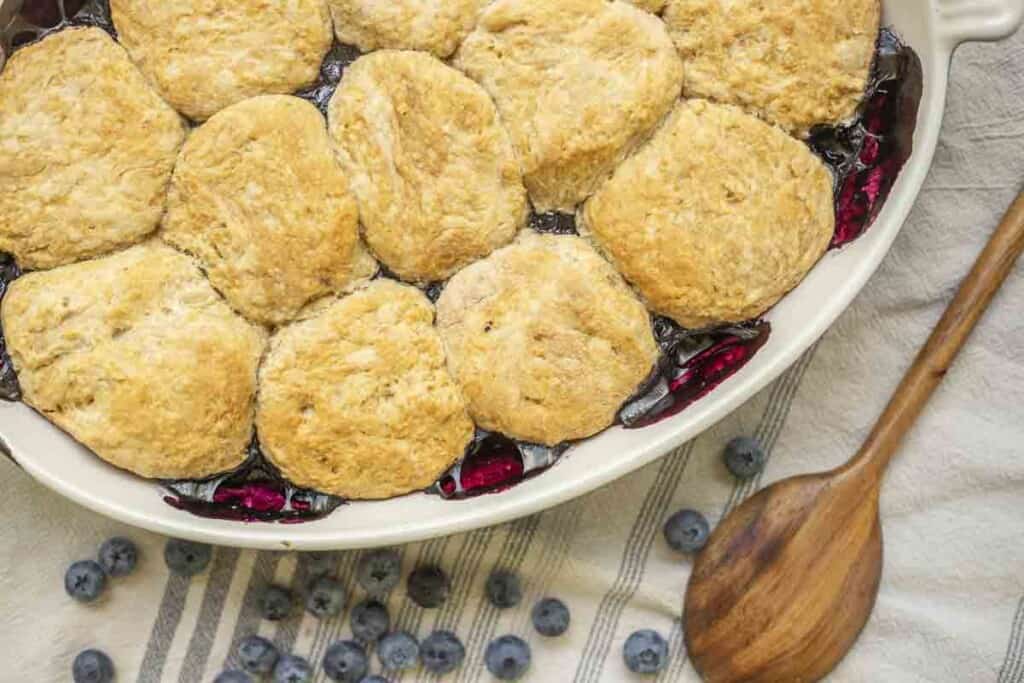 sourdough blueberry cobbler in a white baking dish on a towel. Blueberries and a wooden spoon are on the towel