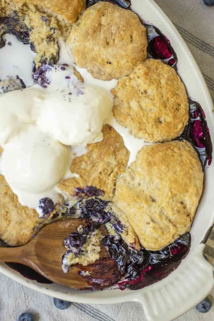 overhead photo of sourdough blueberry cobbler topped with ice cream with a wooden spoon in the cobbler