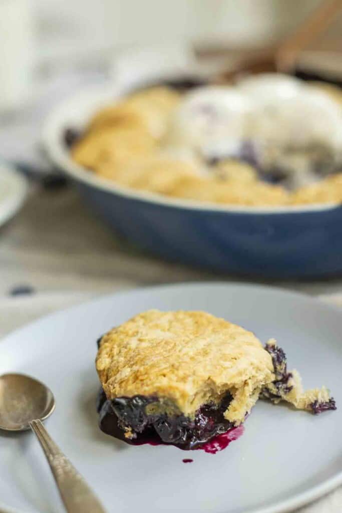 blueberry cobbler on a gray plate with a spoon with more cobbler in a blue baking dish in the background