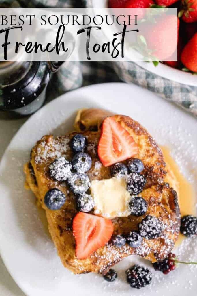 overhead photo of sourdough French toast topped w with blueberries, sliced strawberries, a pat of butter, maple syrup and powdered sugar on a white plate with a bowl of strawberries in the back