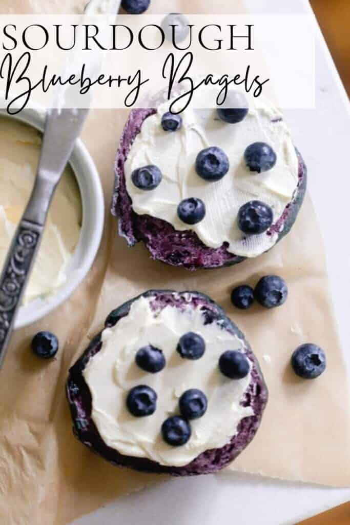 overhead photo of a sourdough blueberry bagel sliced in half and topped with cream cheese topped with blueberries. The bagels are on parchment paper with a cream cheese in a white container and topped with a knife