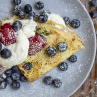 Overhead photo of one sourdough crepes folded with a whipped cream filling and topped with more whipped cream strawberries, blueberries and powdered sugar on a gray plate