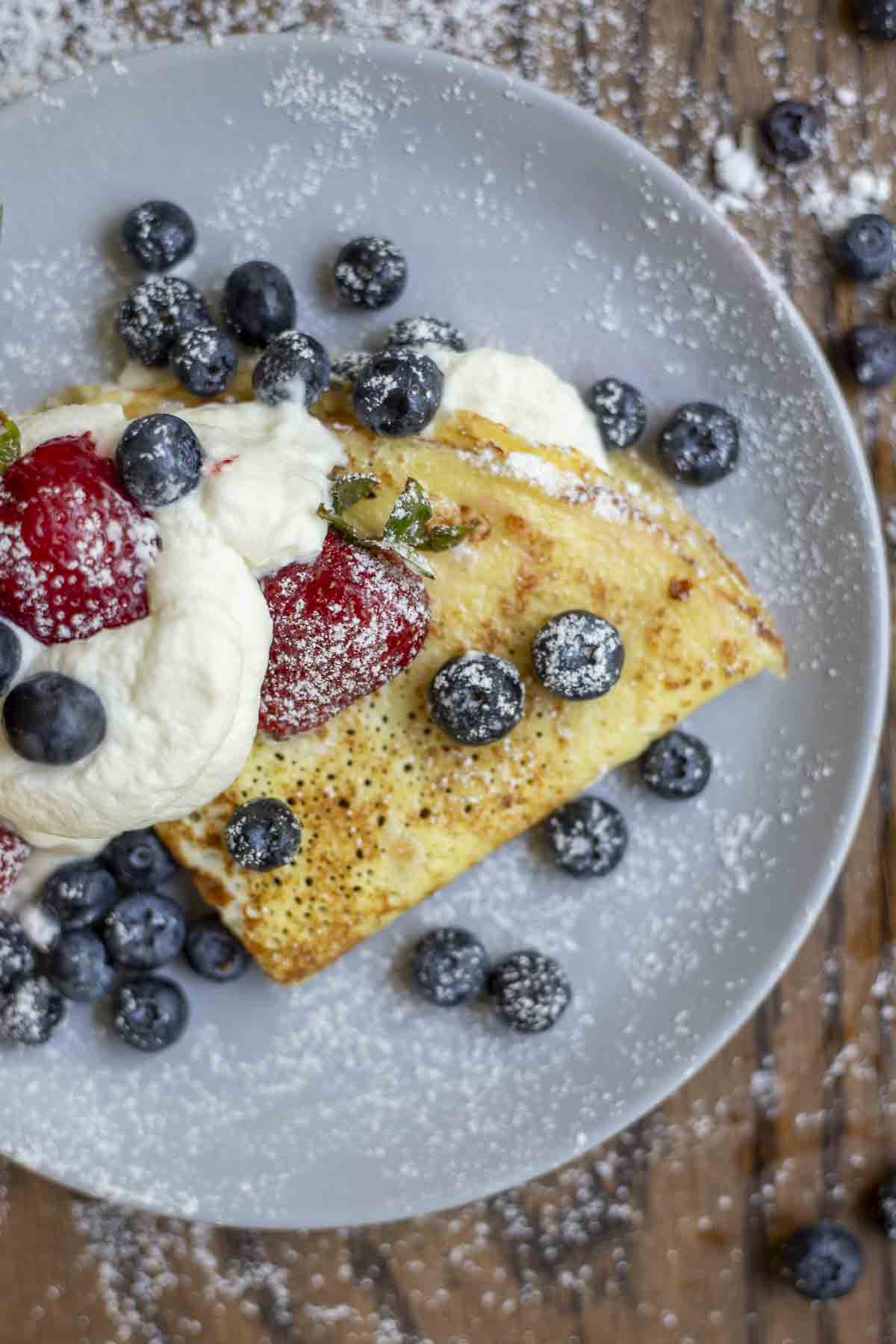 Overhead photo of one sourdough crepes folded with a whipped cream filling and topped with more whipped cream strawberries, blueberries and powdered sugar on a gray plate