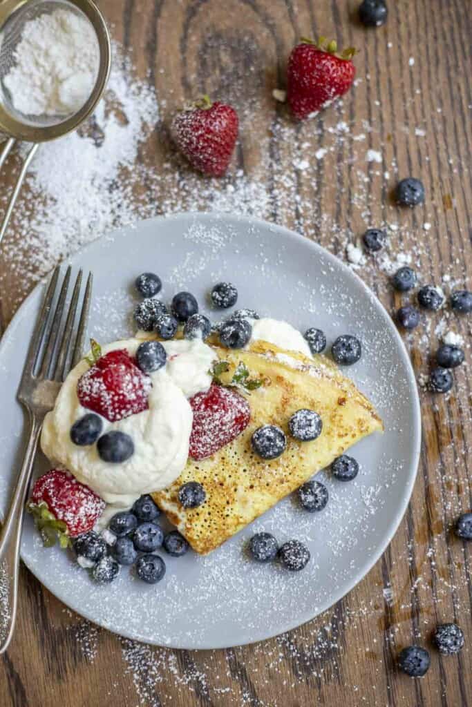 gray plate with one sourdough crepe folded with a bunch of whipped cream topped with blueberries and strawberries. The plate is surrounded by blueberries, strawberries, and a sifter with powdered sugar