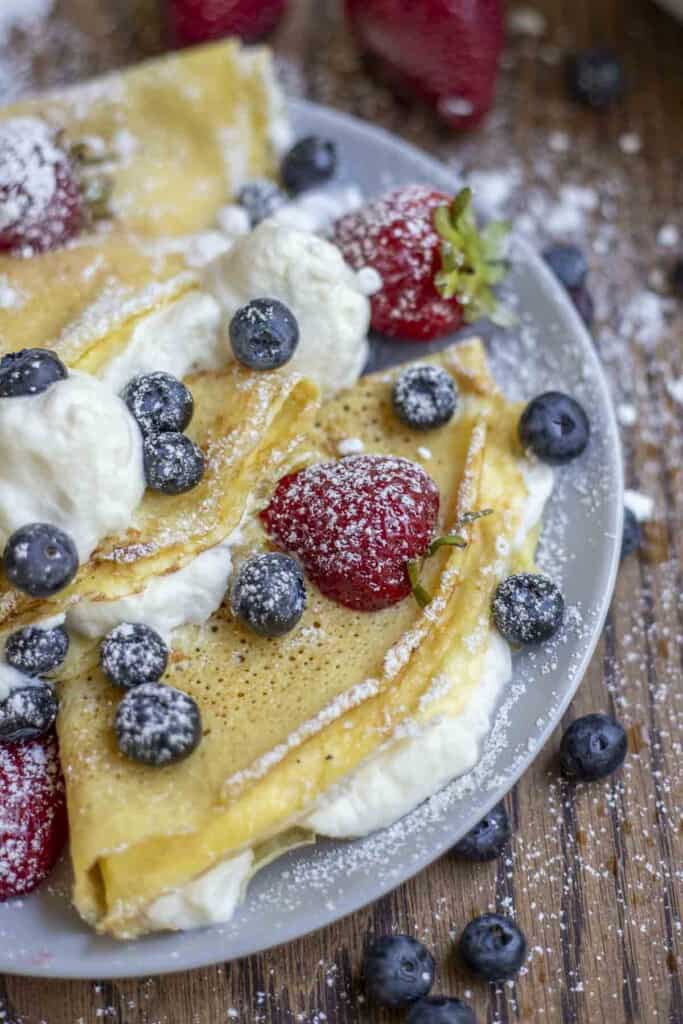 side view of three sourdough crepes with whipped cream filling, blueberries and strawberries on a gray plate on a wood table