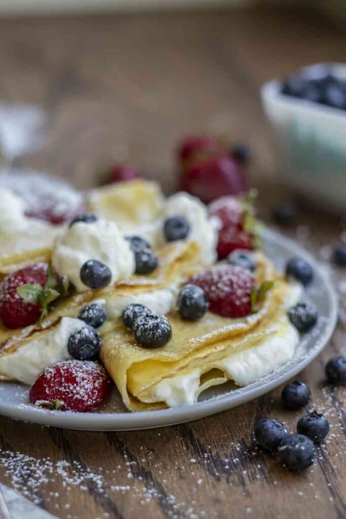 side view of a gray plate with three folded sourdough crepes with whipped cream, strawberries, and blueberries topped on the crepes. The plate is on a wood table with blueberries and strawberries scattered around